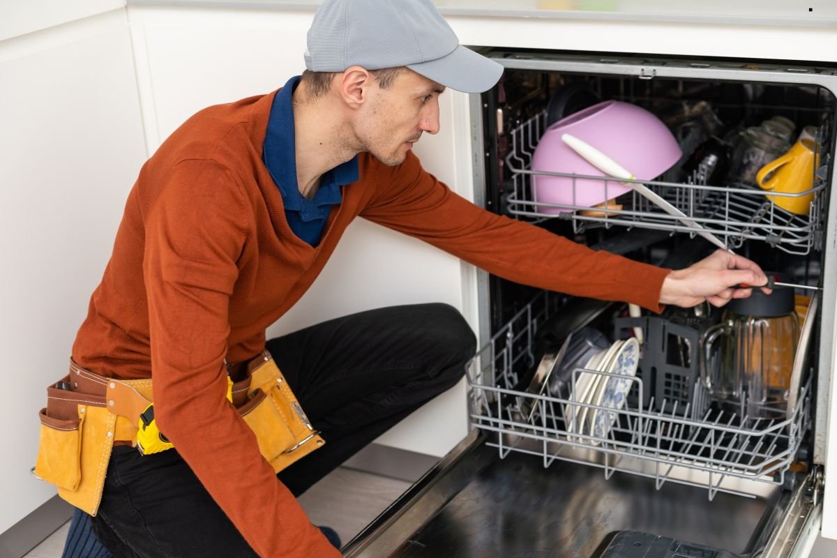 A person in a brown sweater and cap kneels beside a dishwasher, adjusting items inside the open appliance.