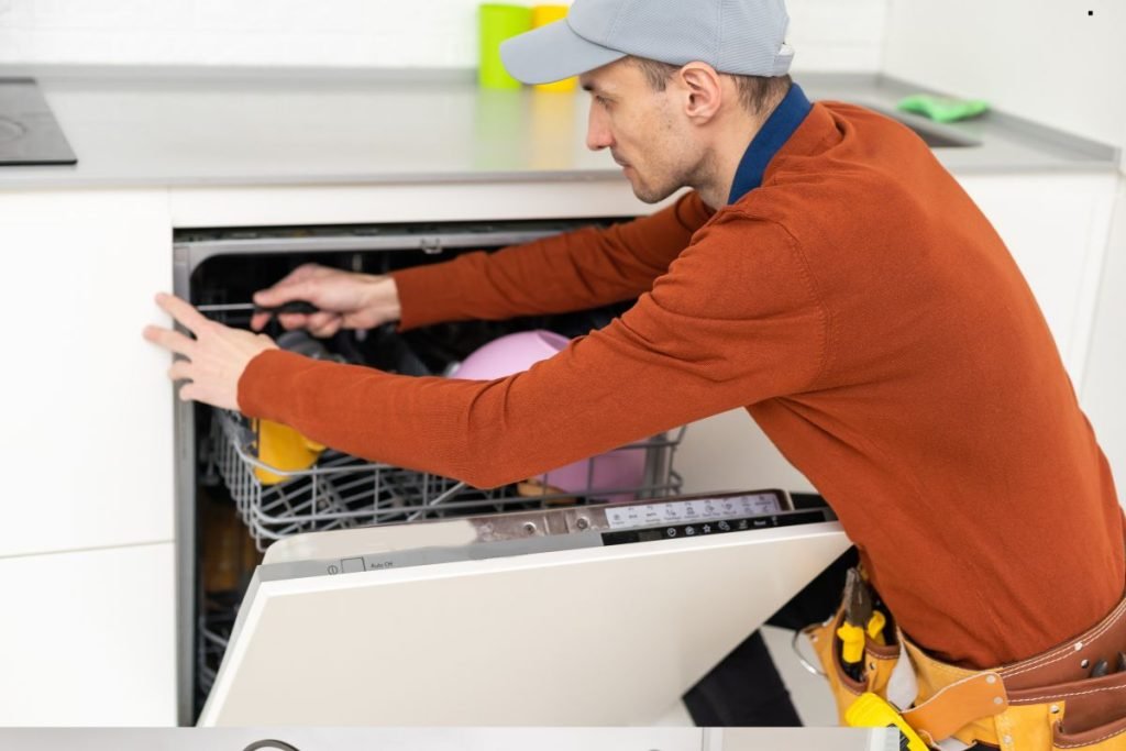 A man in a red sweater and cap repairs a dishwasher in a modern kitchen. He appears focused, with tools visible in his belt. Bright and organized scene.